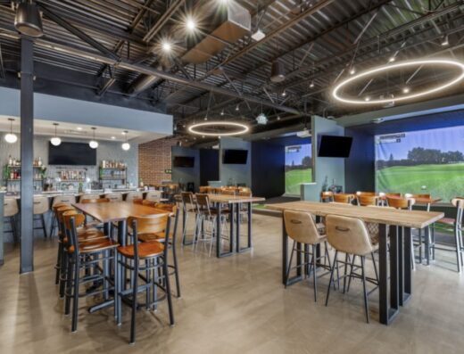 Modern indoor bar and lounge with high tables and chairs, ring-style ceiling lights, a bar area in the back, and large golf simulator screens on the right side showing golf courses.