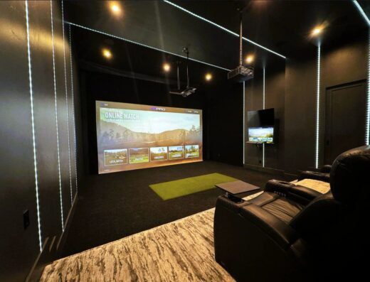 A modern home theater room with a large screen displaying a golf game, leather recliners, a small green golf mat on the floor, wall-mounted TV, dark walls, and LED strip lighting accents.