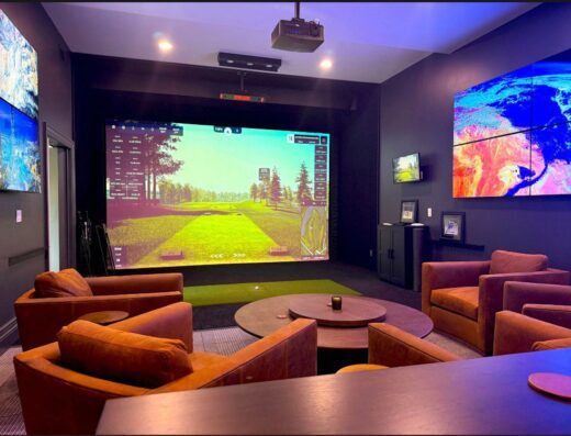 A modern room with brown armchairs arranged around a round coffee table, facing a large golf simulator screen. The walls display colorful landscape images on multiple monitors. The space is softly lit and contemporary.