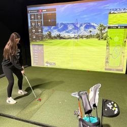 A woman prepares to swing a golf club at an indoor golf simulator, with a digital display of a golf course projected on the wall in front of her. A golf bag and clubs are visible in the foreground.