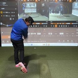 A person in a blue shirt and black pants swings a golf club indoors, with golf simulation data and swing analysis displayed on a large screen in front of them.