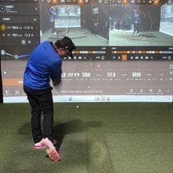 A person in a blue shirt and black pants swings a golf club indoors, with golf simulation data and swing analysis displayed on a large screen in front of them.