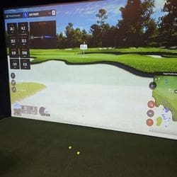 A golf simulator screen displays a virtual golf course with distances, stats, and a green surrounded by sand traps; two yellow golf balls are on the green mat in front of the screen.