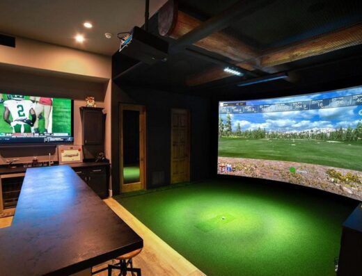 A home entertainment room with a large TV showing a football game on the left and a curved screen golf simulator on the right, with artificial turf flooring and dark modern decor.