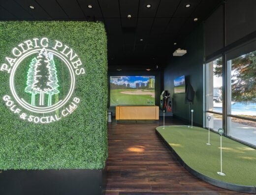 Indoor golf facility with a putting green, golf simulator screen, and a wall covered in greenery featuring a neon sign that reads Pacific Pines Golf & Social Club. Large windows provide natural light.