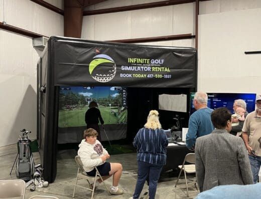 People gather around an indoor golf simulator booth labeled INFINITE GOLF SIMULATOR RENTAL in a large room. Some are seated, while others stand near the entrance or watch the screen inside.