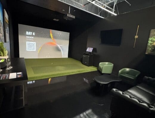 Modern indoor golf simulator room with a large screen, artificial turf, lounge seating, a TV, a sword on the wall, and glossy black floors under an open ceiling with exposed pipes and wires.