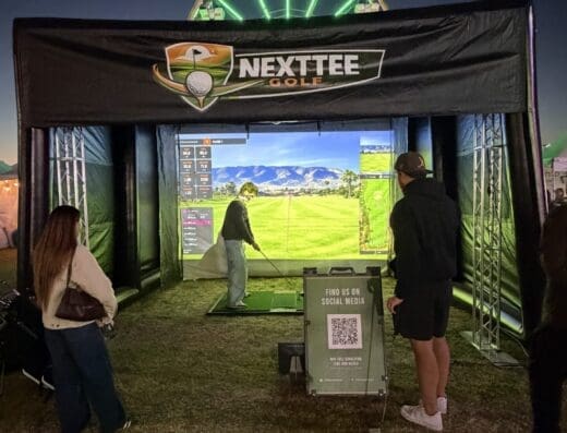 People play virtual golf at night in an outdoor tent with a NEXTTEE GOLF sign. A screen shows a golf course, and a Ferris wheel glows with green lights behind the tent.