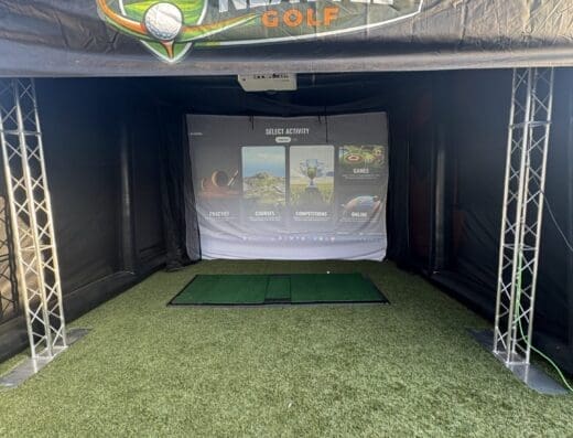 A golf simulator setup under a tent labeled NEXT TEE GOLF, featuring a projection screen displaying activity options, artificial turf, golf mats, and a launch monitor on the ground.