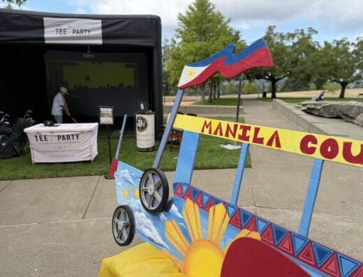A colorful Manila-themed cart with sun and flag designs stands near a park. In the background, a person in a mask plays a golf simulator under a black tent labeled Tee Party. Trees and walkways are visible.